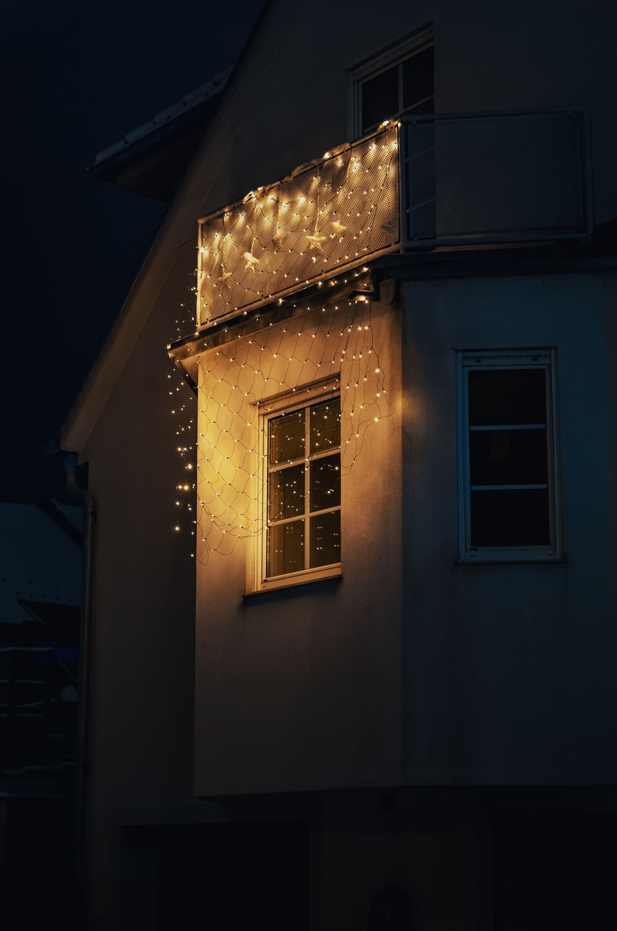 The side of a house at night with a balcony rail wrapped in warm white lights above a glowing window
