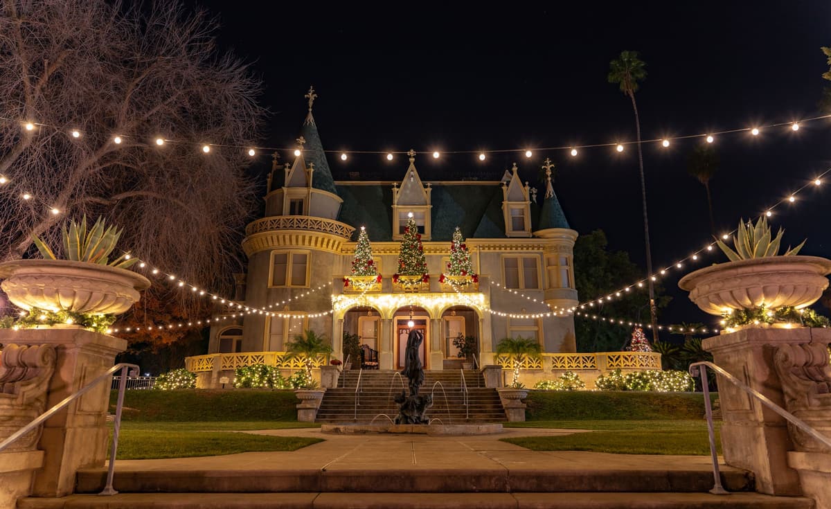 A historic hillside estate at night, its arched porch outlined in warm white lights, with swags of bulb lights over the garden steps and tall palms behind