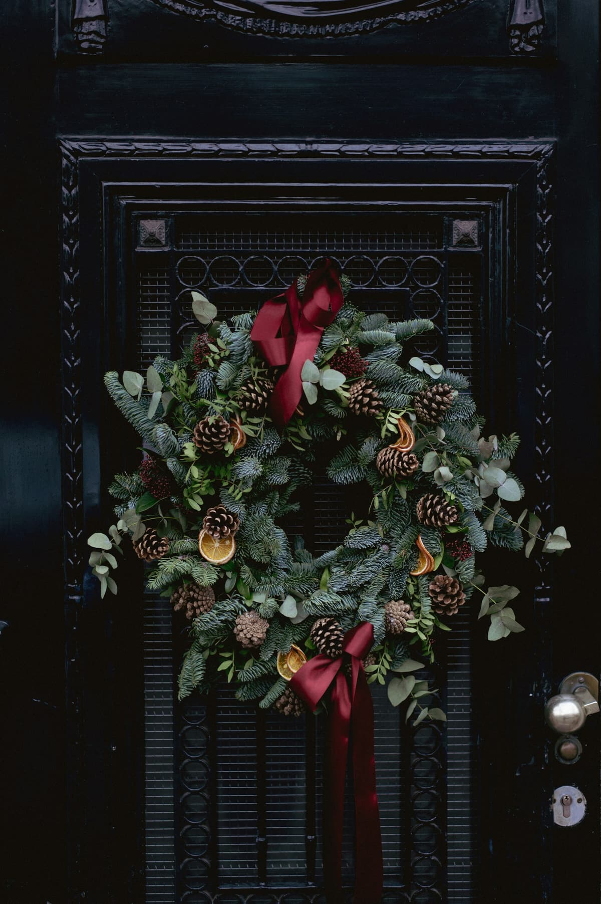 A wreath of eucalyptus, pine and pinecones with a red ribbon on a dark paneled front door