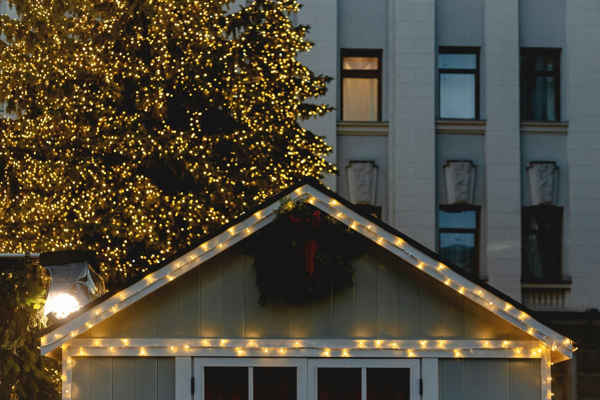 Gabled building trimmed with evenly spaced warm white bulbs and a wreath, in front of a tall tree covered in lights